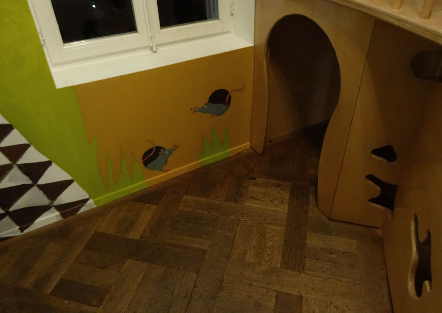 Wandmalerei mit zwei grauen Mäusen; eine verlässt und eine betritt ein schwarzes Loch auf gelbem Hintergrund, umgeben von grünen Gräsern.