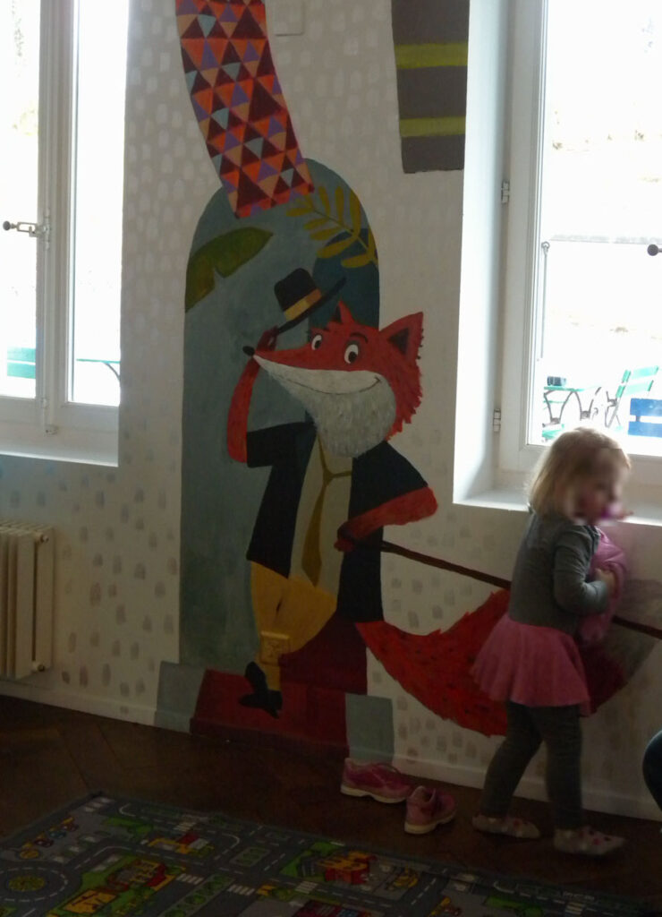 Wandgemälde in einem Kinderzimmer: Ein stilisierter roter Fuchs mit Gehstock und Zylinder steht in einer Art Türbogen. Der Fuchs trägt ein Jackett, gelbe Hose und eine Krawatte und hebt grüßend den Hut. Der Hintergrund zeigt stilisierte Pflanzenmotive und Muster. Am Boden liegt ein bunter Spielteppich mit Straßenmotiv sowie ein Paar rosa Kinderschuhe. Die Szene wirkt freundlich, verspielt und leicht nostalgisch.
